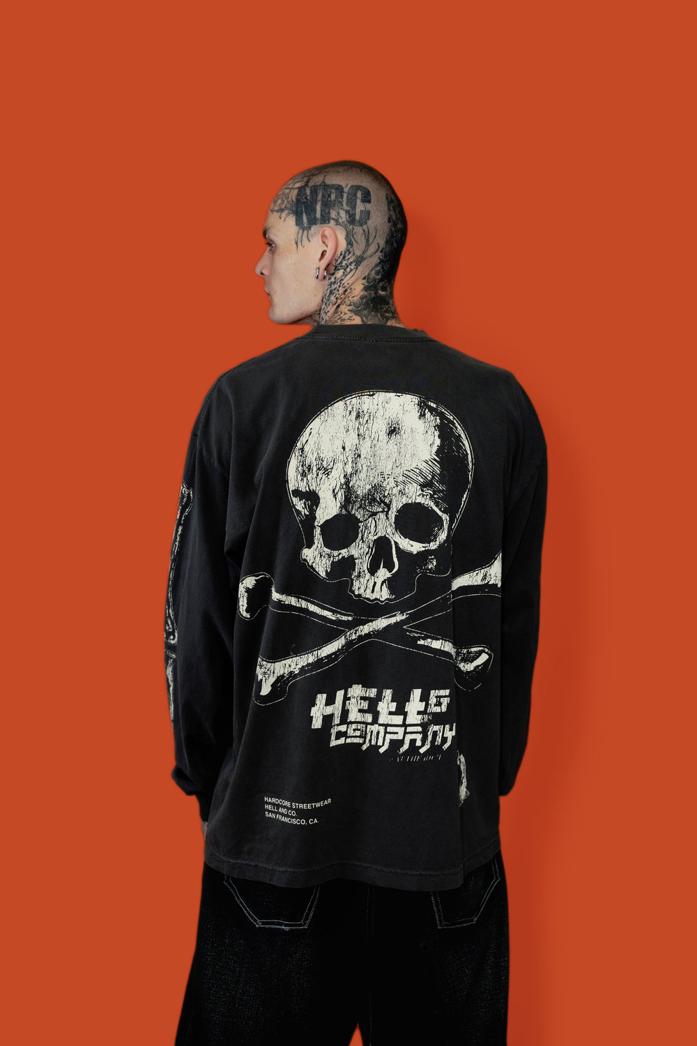 A man with tattoos is shown from the back wearing a black long-sleeve shirt with a large white skull and crossbones graphic and the text 'HELL OR BURONY' on the back, set against a solid orange background.