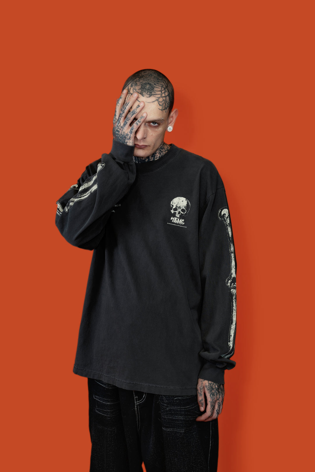 A tattooed man posing in front of a solid orange background, wearing a black long-sleeve graphic t-shirt with a white skull and 'AMC' logo on the chest, paired with black jeans.