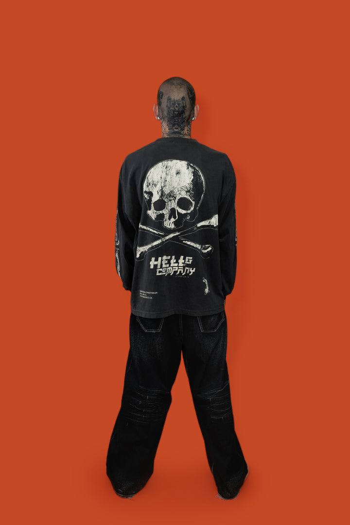 A man stands with his back to the camera wearing a black long-sleeve graphic shirt and black pants against a solid orange background. The shirt features a large white skull and crossbones graphic on the back with the text 'HELLO COMPANY' printed below.