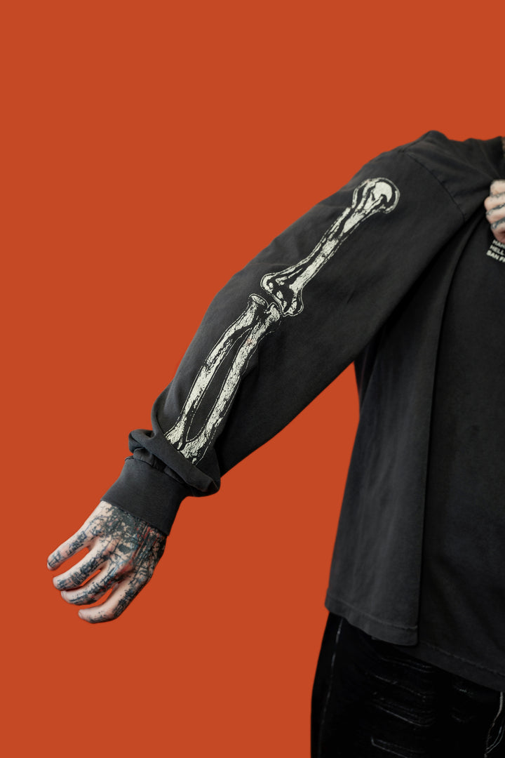 A person wearing a black long-sleeve shirt with a large white skeleton arm graphic, shown against a solid orange background. The person's tattooed hand is visible.
