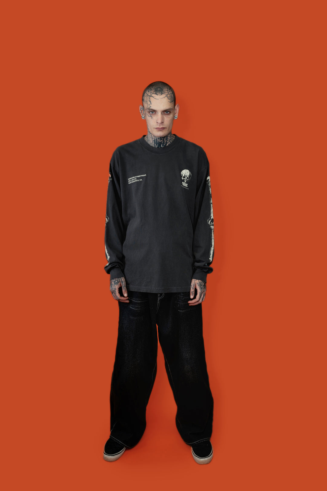A tattooed model stands against a solid orange background wearing a washed black long-sleeve graphic shirt, black baggy pants, and black sneakers.