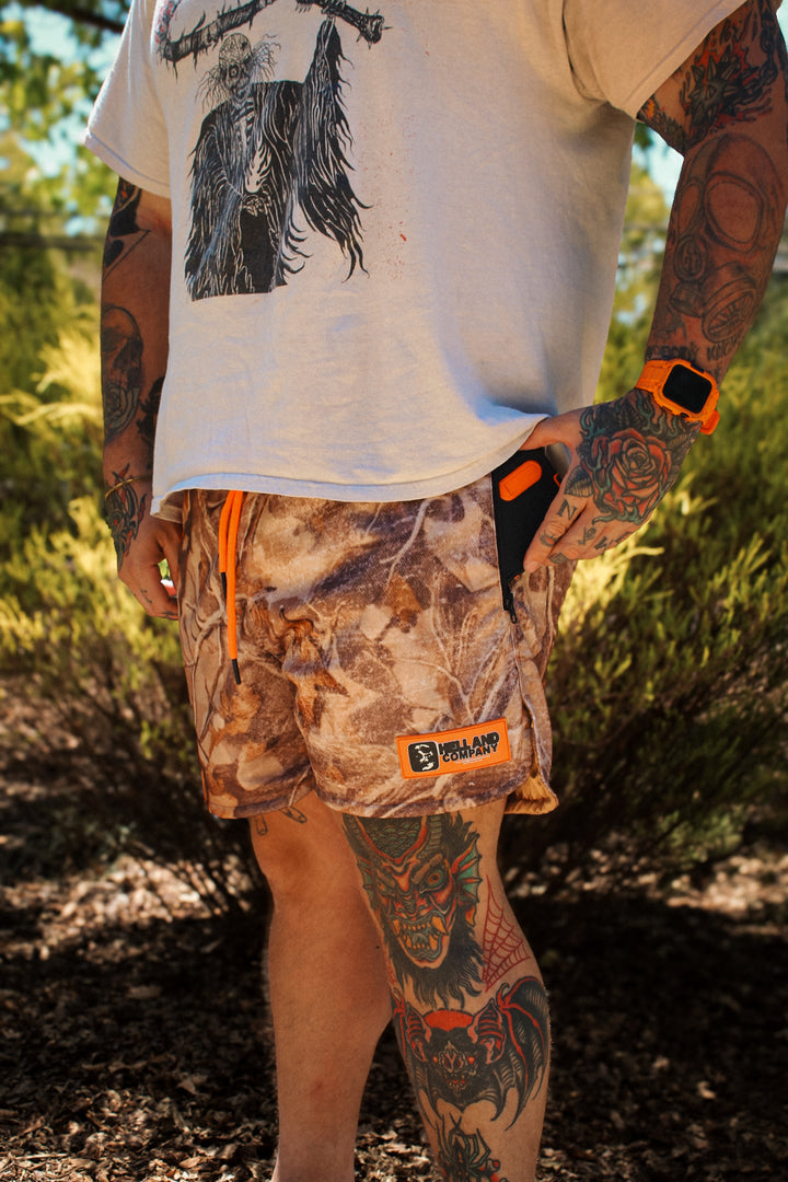 Person wearing a white t-shirt with a graphic design and camouflage shorts with orange accents in a forest setting.
