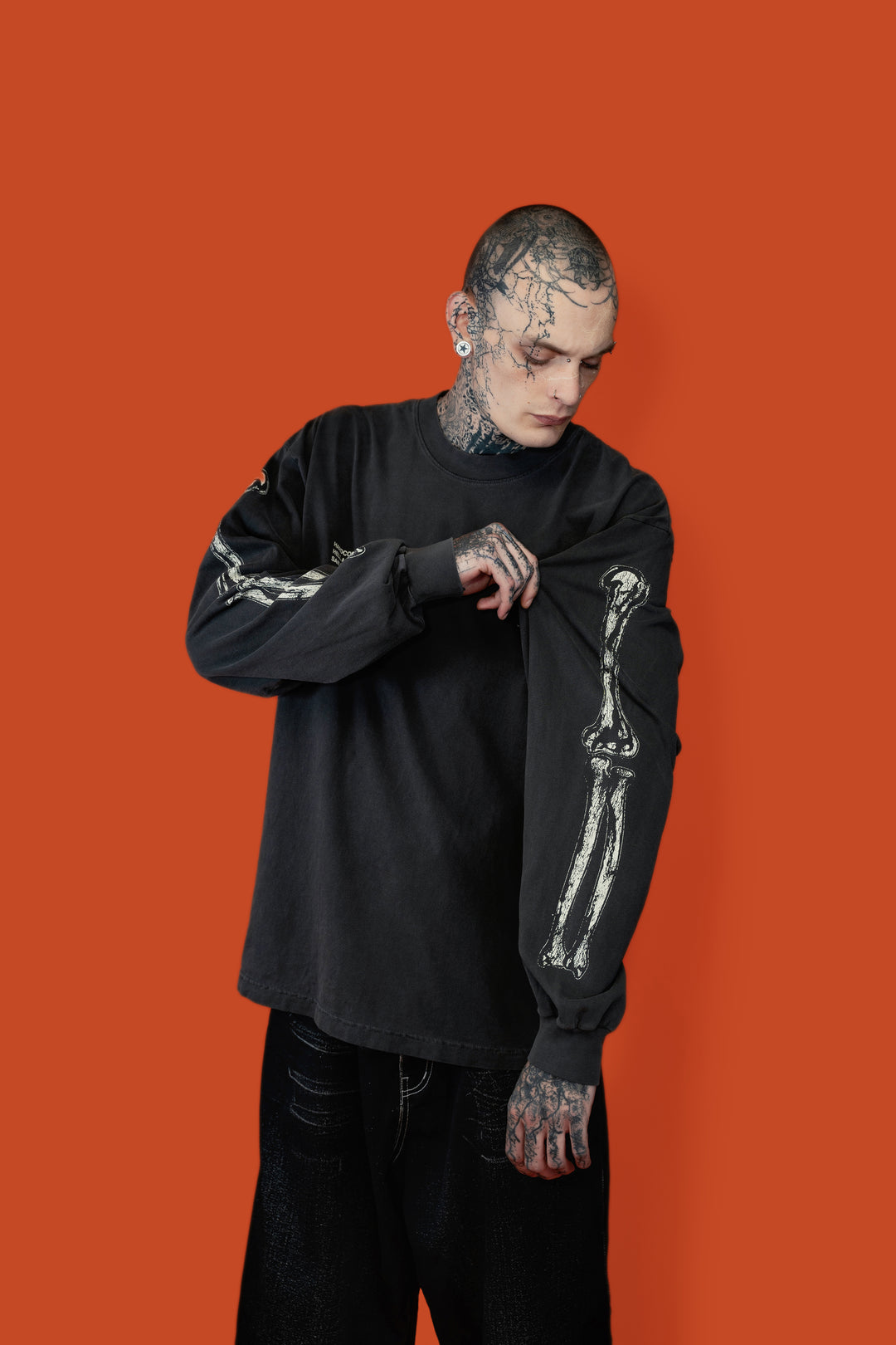 A tattooed man wearing a black long-sleeve crewneck shirt with a white skeleton graphic on the sleeve, posed against a solid orange background.