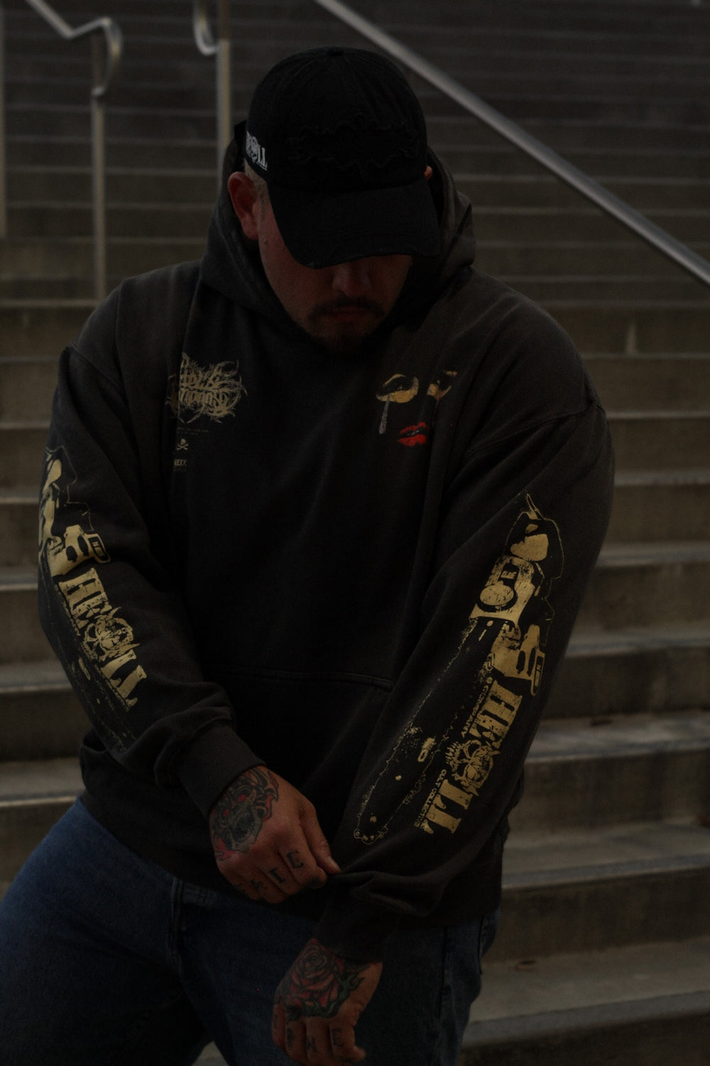 A man wearing a black hoodie with gold graphic prints on the sleeves, blue jeans, and a black baseball cap, sitting on concrete stairs.