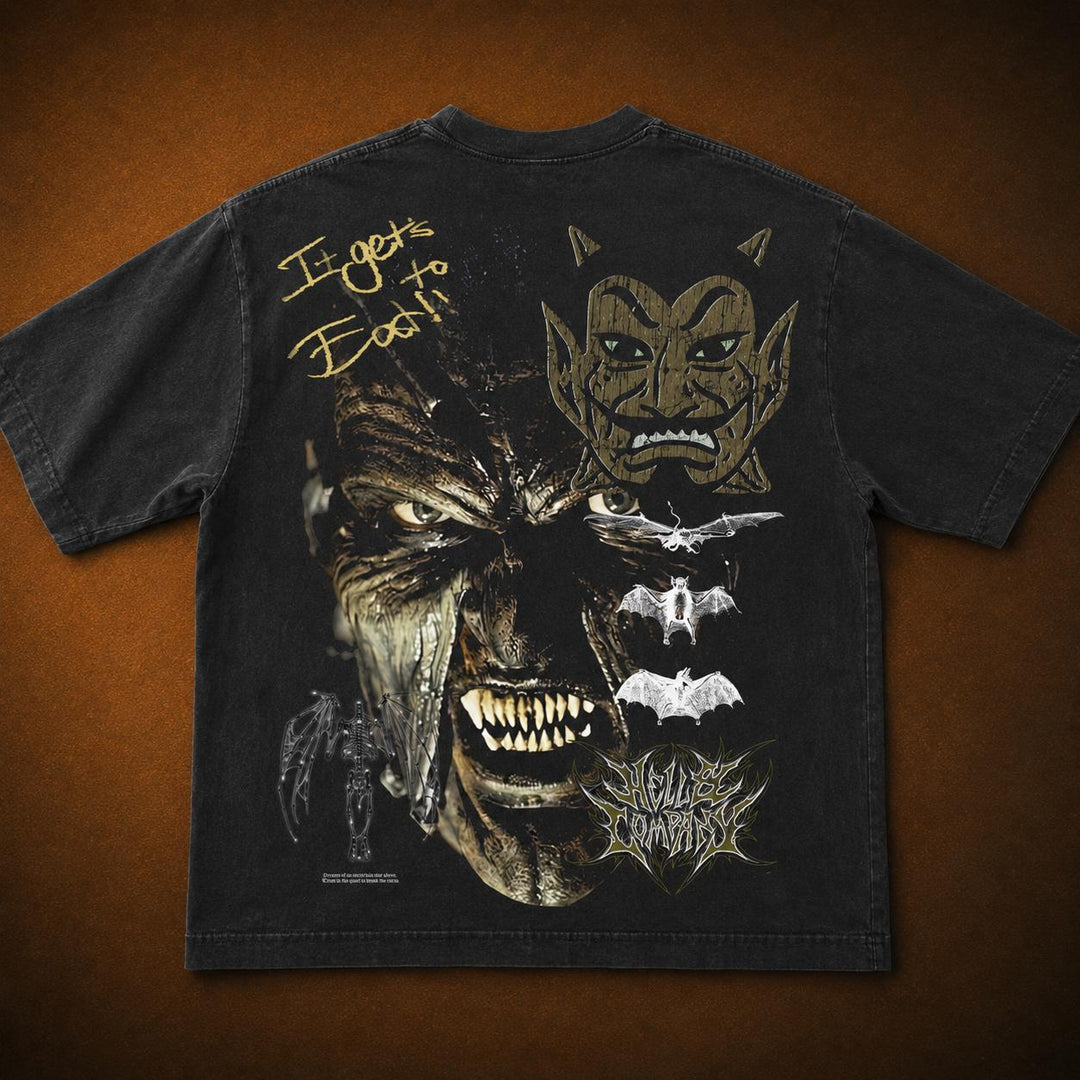 A black graphic t-shirt laid flat against a textured brown background, showing the back side with a large detailed print featuring a demonic face, bats, a spider, and gold text reading 'It Gets Better!'.